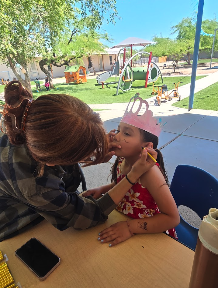 A teacher painting a small design on a child’s face at an outdoor event