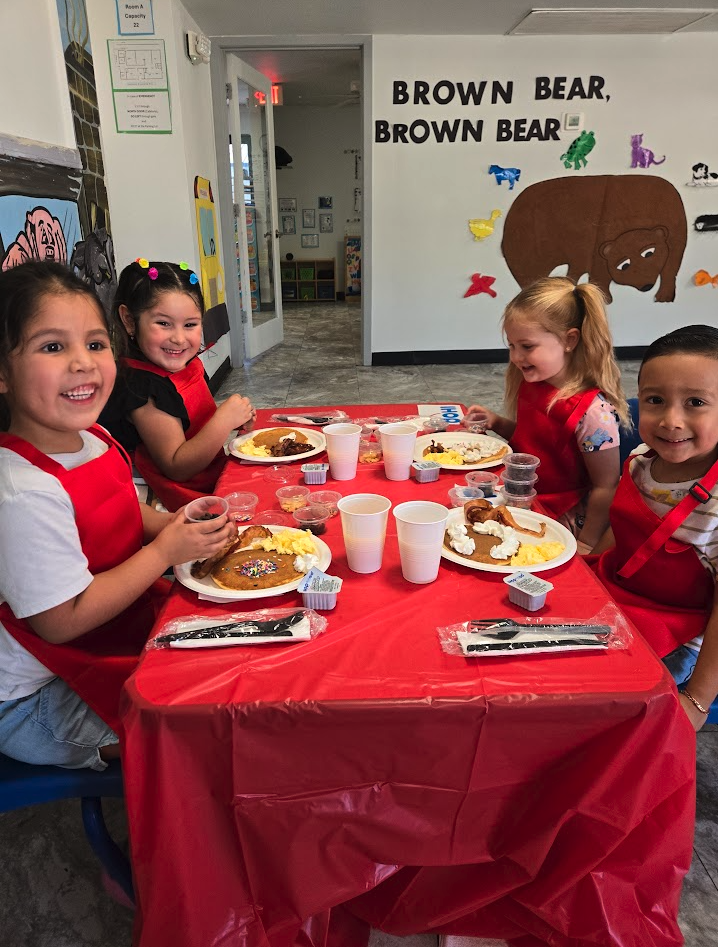Four smiling children in red aprons at a pancake breakfast table